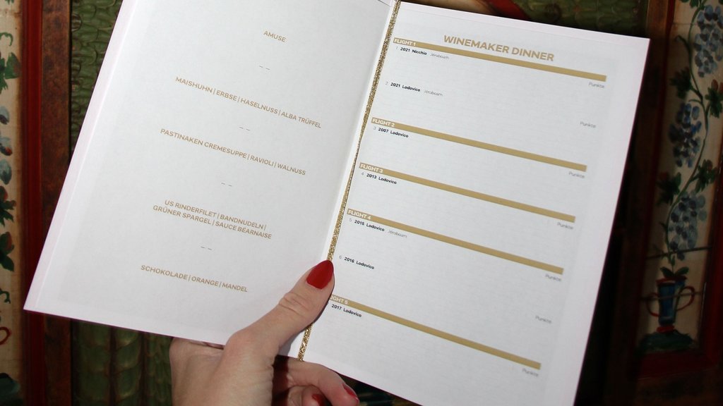 Hand hält Menükarte mit Speisen und Winemaker Dinner Weinauswahl vor dekorativem Holzpaneel