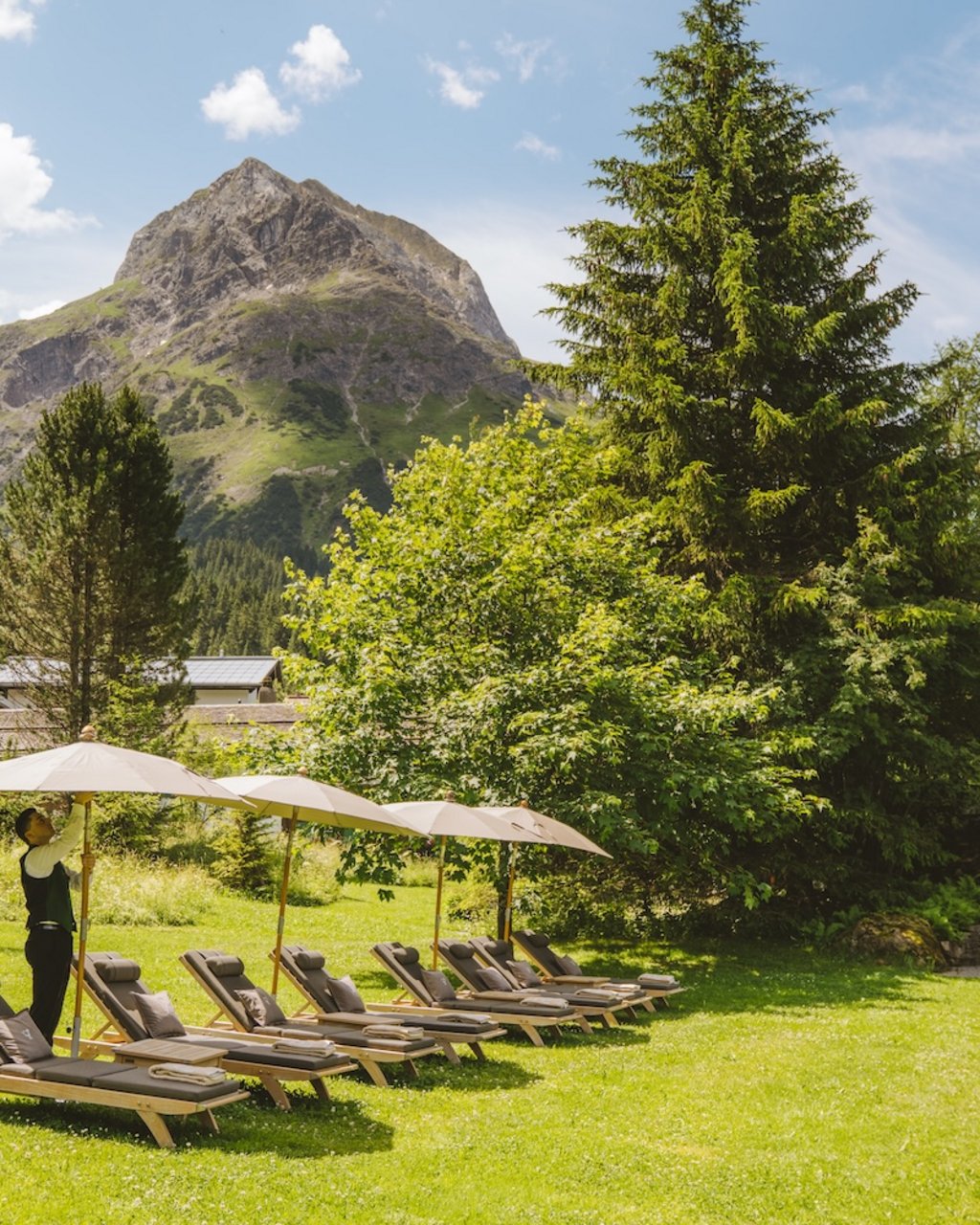 Man sets up umbrellas by lounge chairs in a garden with mountain backdrop