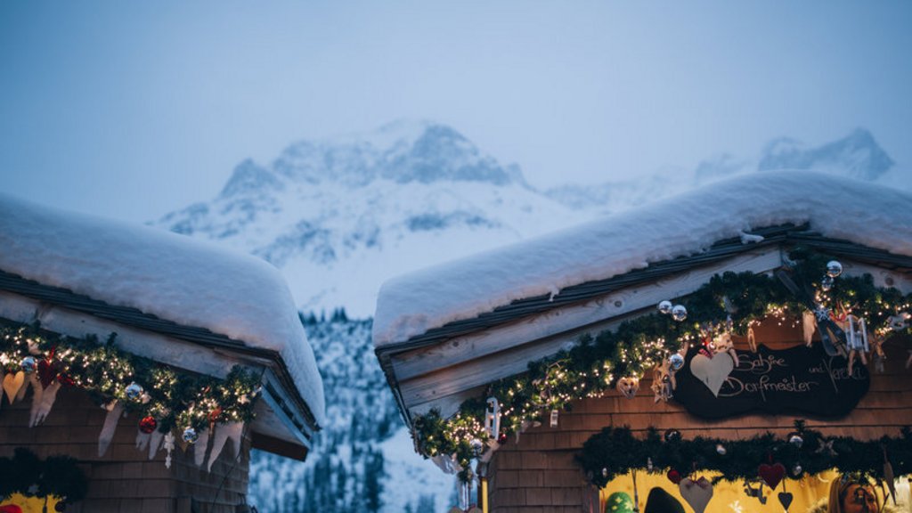 Adventzauber Schneebedeckte Hütten mit Weihnachtsbeleuchtung vor verschneiten Bergen