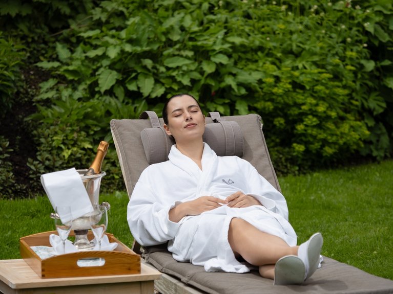 Woman relaxing on lounge chair in robe with champagne and glasses in garden