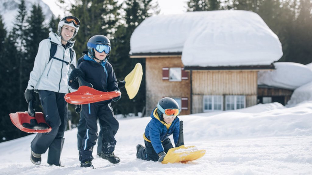 Frau und zwei Kinder mit Schleifschaufeln spielen im Schnee vor verschneiter Hütte
