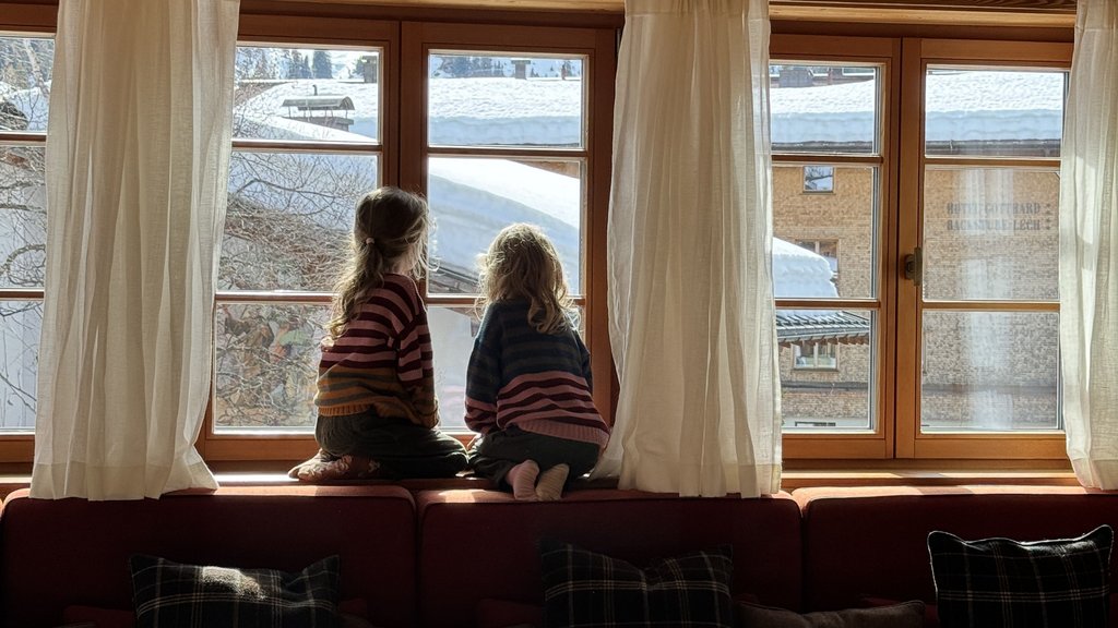 Zwei Kinder sitzen auf einem Fenstersims und blicken nach draußen auf schneebedeckte Dächer