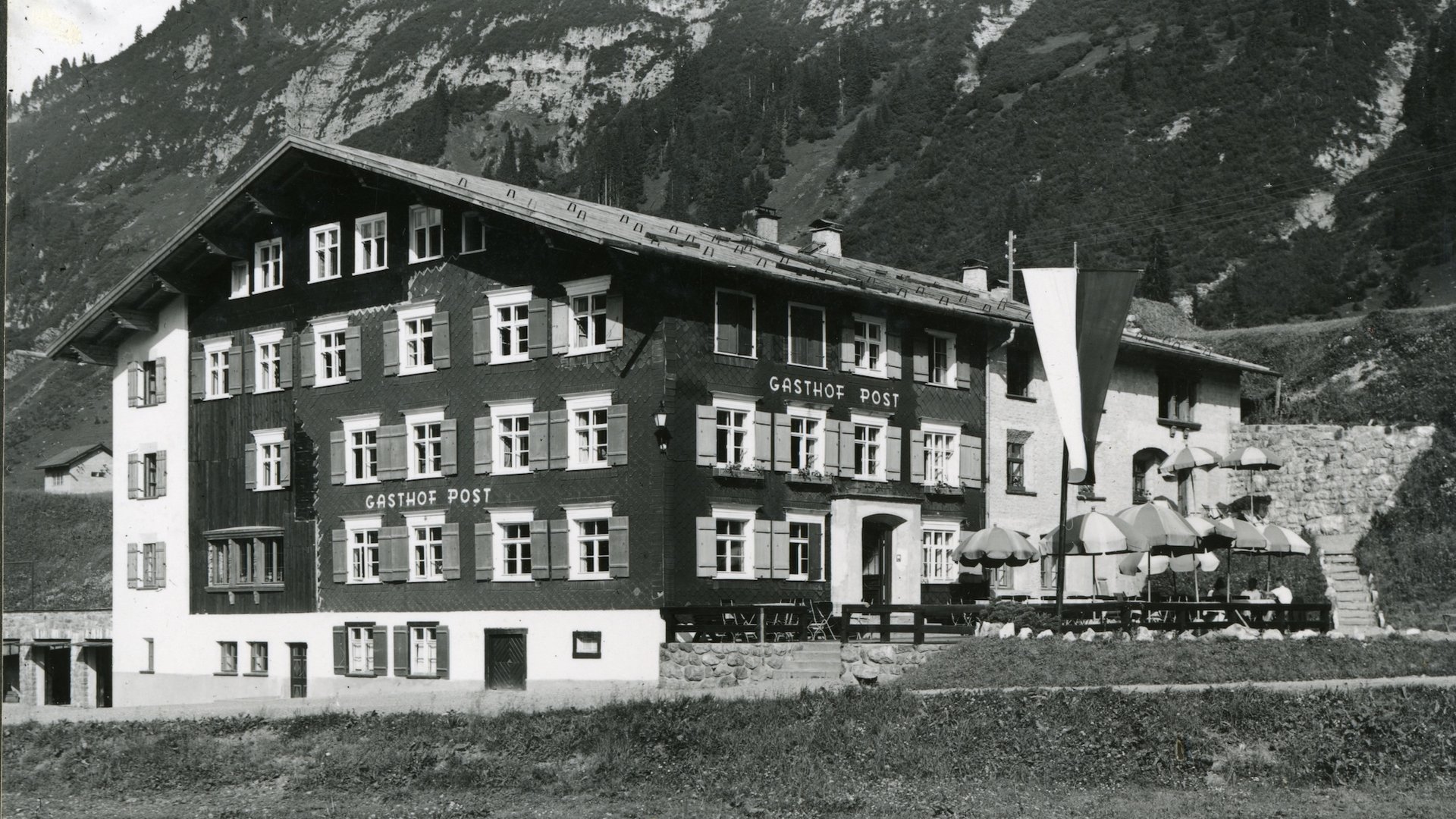 Ein Haus mit Geschichte. Eine Geschichte mit Zukunft. Historisches Gasthof Post mit Sonnenschirmen vor alpiner Bergkulisse