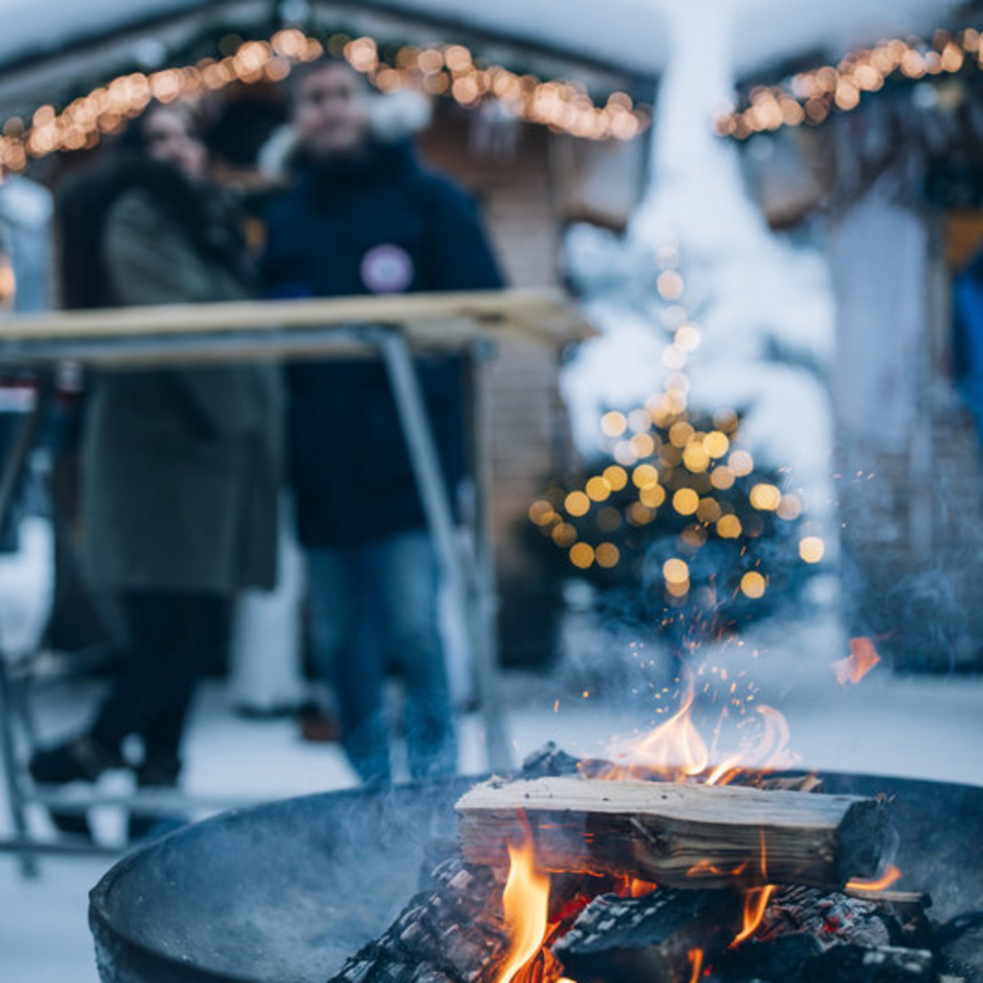Adventzauber Holzfeuer in Schale mit unscharfen Personen und Lichterkette im verschneiten Hintergrund